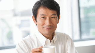 白いシャツを着た男性がコーヒーカップを持ち、カメラに向かって微笑んでいる。